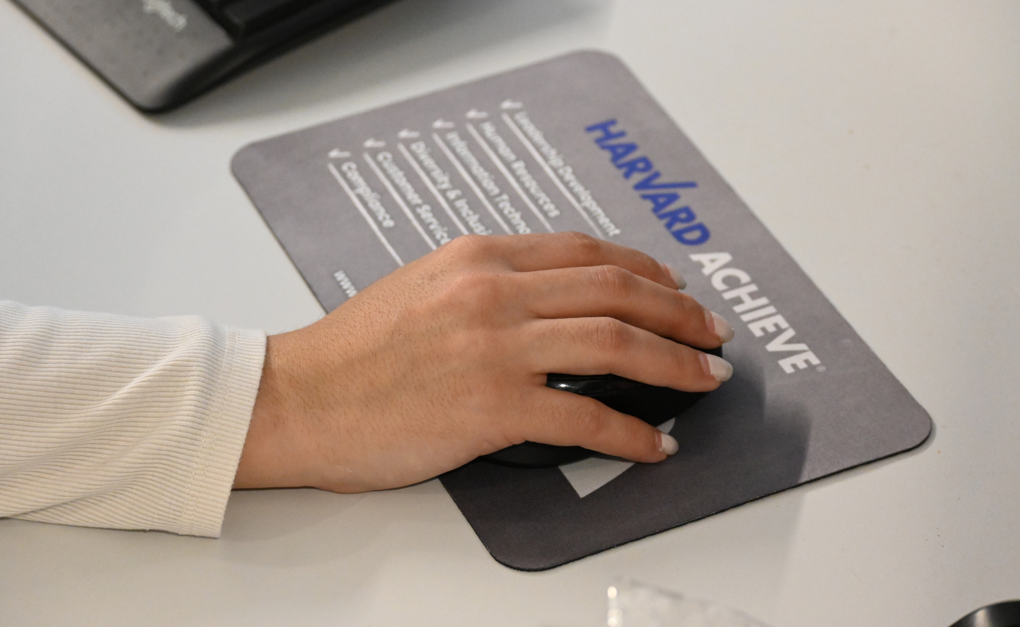 A hand using a computer mouse on a mousepad labeled "HARVARD ACHIEVE" with a checklist of goals.