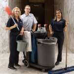 Three janitors with cleaning supplies stand together in a marble-floored building hallway.