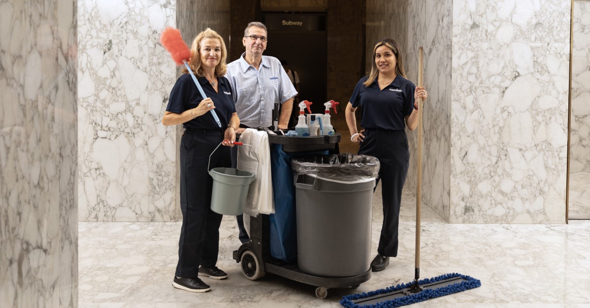 Three janitors with cleaning supplies stand together in a marble-floored building hallway.