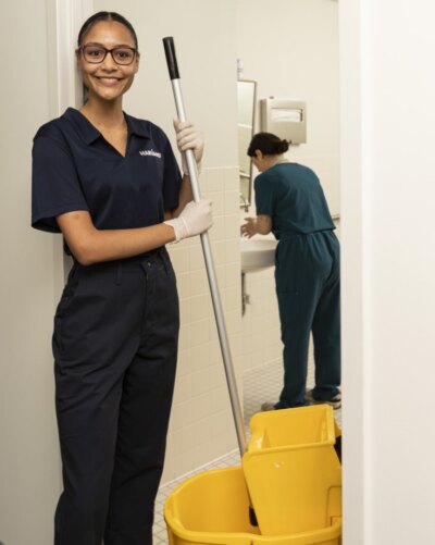 Two people cleaning a restroom, one holding a mop and the other cleaning a sink.