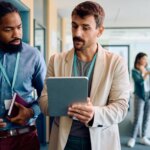 Two men in an office discussing something on a tablet while others talk in the background.