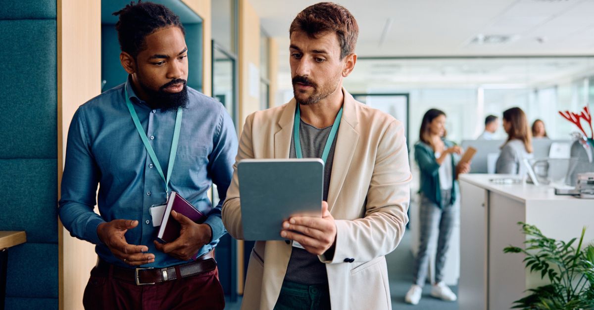 Two men in an office discussing something on a tablet while others talk in the background.