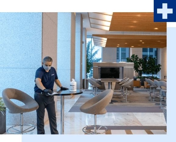 Person cleaning a table in a modern office lobby, wearing a mask and gloves.