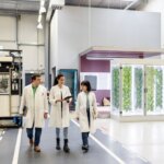 Three researchers in lab coats walking in a modern laboratory space with indoor plants.