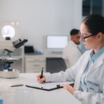 Scientist in a lab coat writing notes at a desk with microscope and computer.