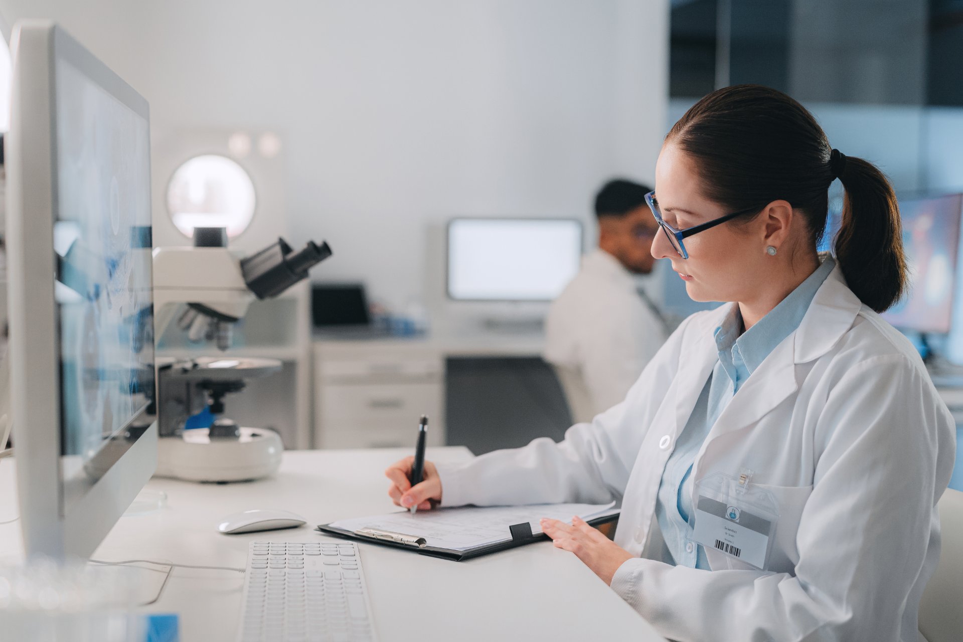 Scientist in a lab coat writing notes at a desk with microscope and computer.